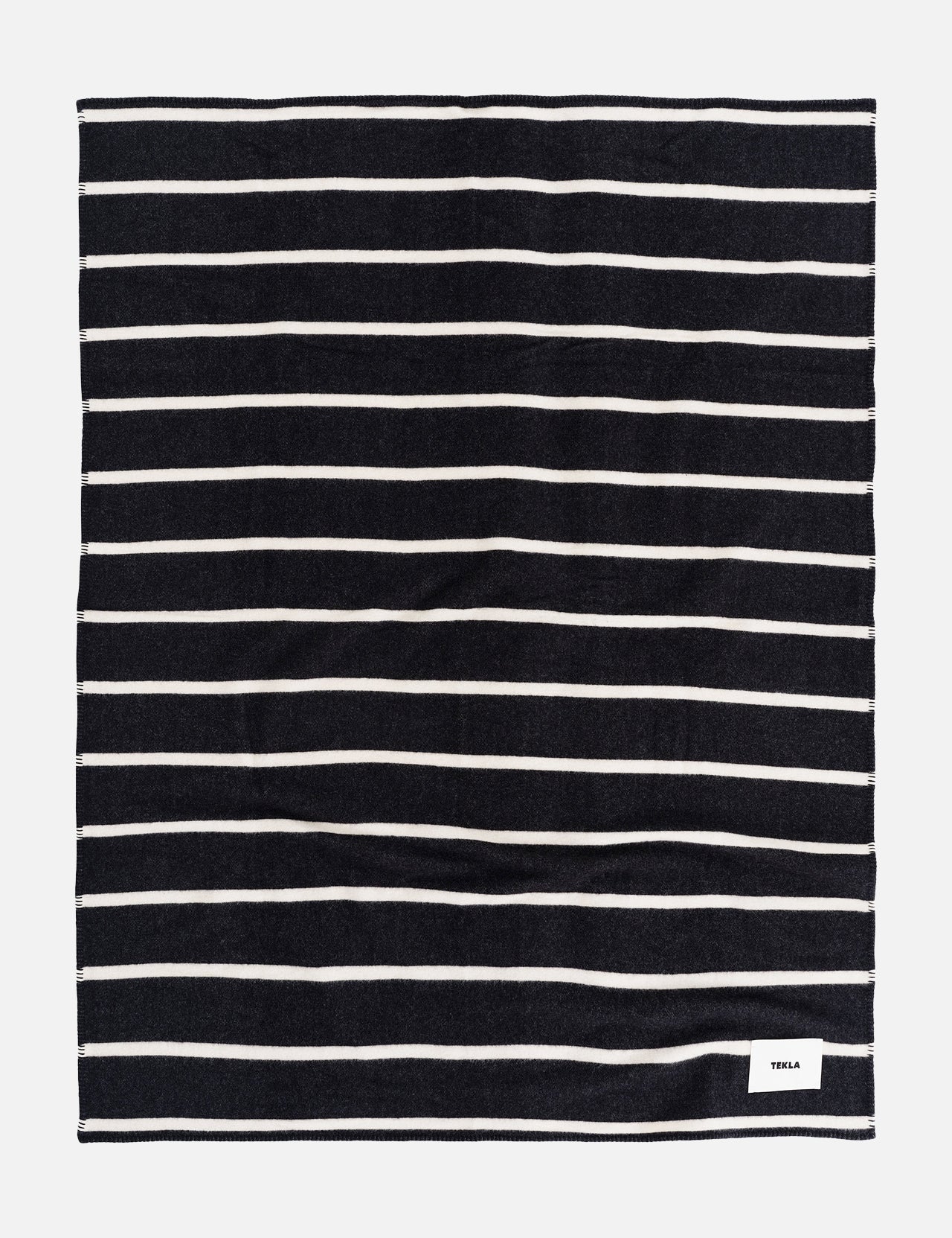A rectangular TEKLA Striped Lambswool Blanket with dark navy and thin white horizontal stripes, featuring a small white logo tag in the bottom corner.