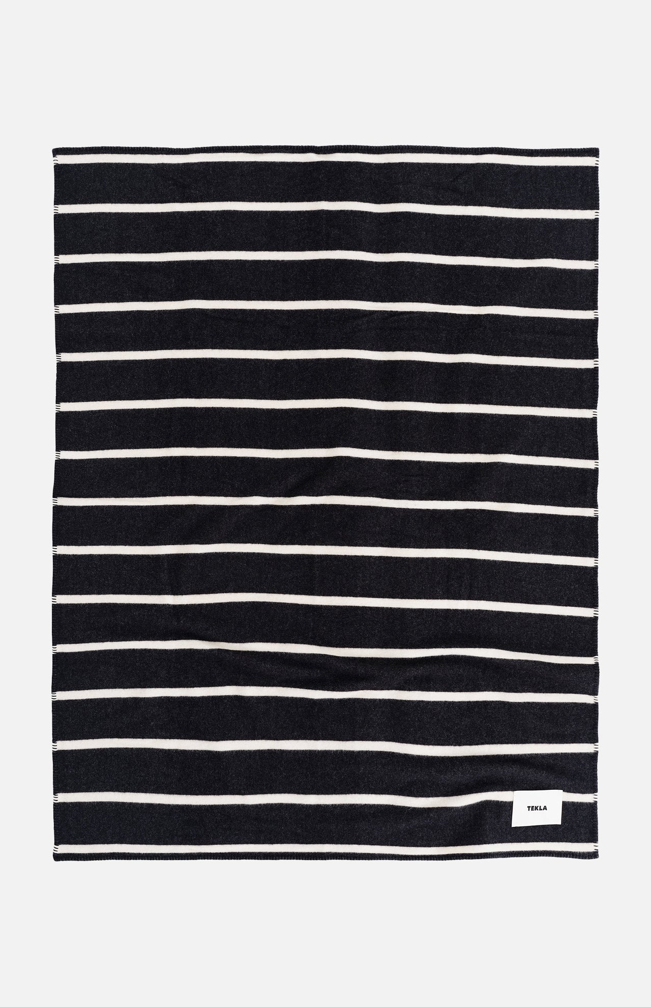 A rectangular TEKLA Striped Lambswool Blanket with dark navy and thin white horizontal stripes, featuring a small white logo tag in the bottom corner.