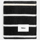 Folded black TEKLA Striped Lambswool Blanket with white horizontal stripes and a central white logo tag.