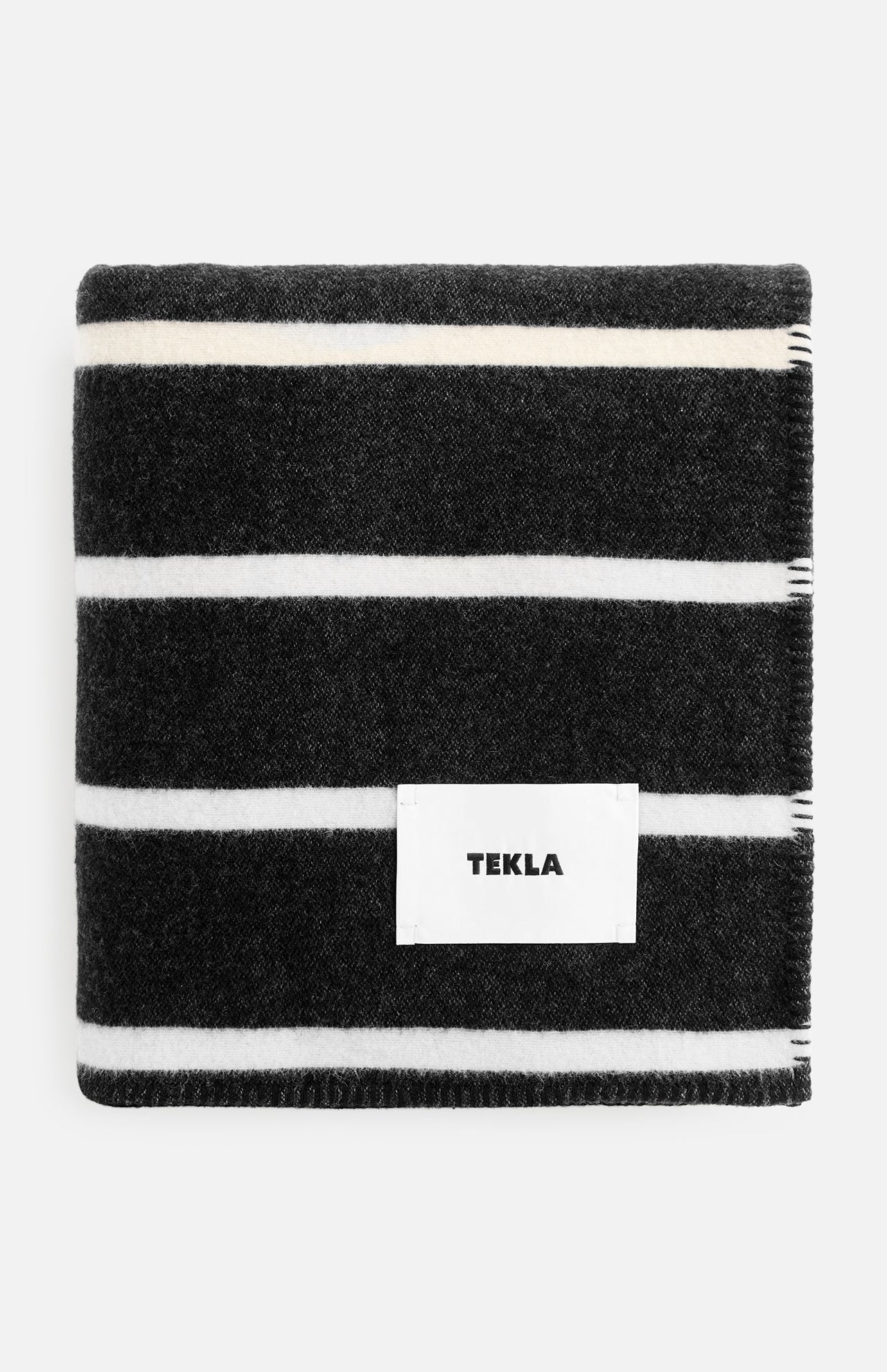 Folded black TEKLA Striped Lambswool Blanket with white horizontal stripes and a central white logo tag.