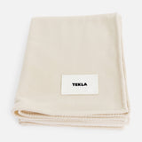 A folded off-white TEKLA Lambswool Blanket with a white label sewn on the front.