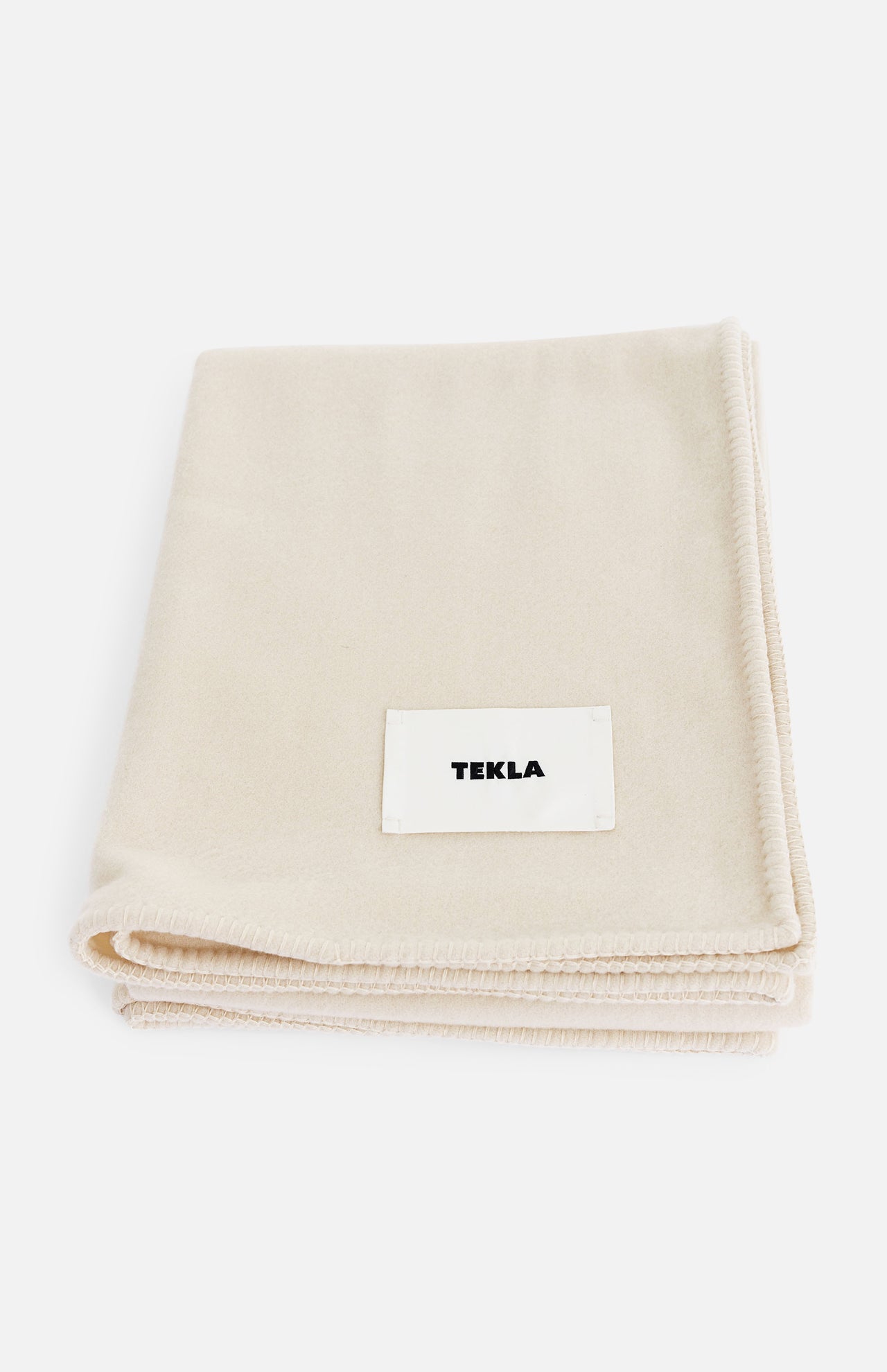 A folded off-white TEKLA Lambswool Blanket with a white label sewn on the front.