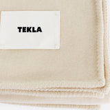 Cream-colored TEKLA Lambswool Blanket with a stitched border and a label.