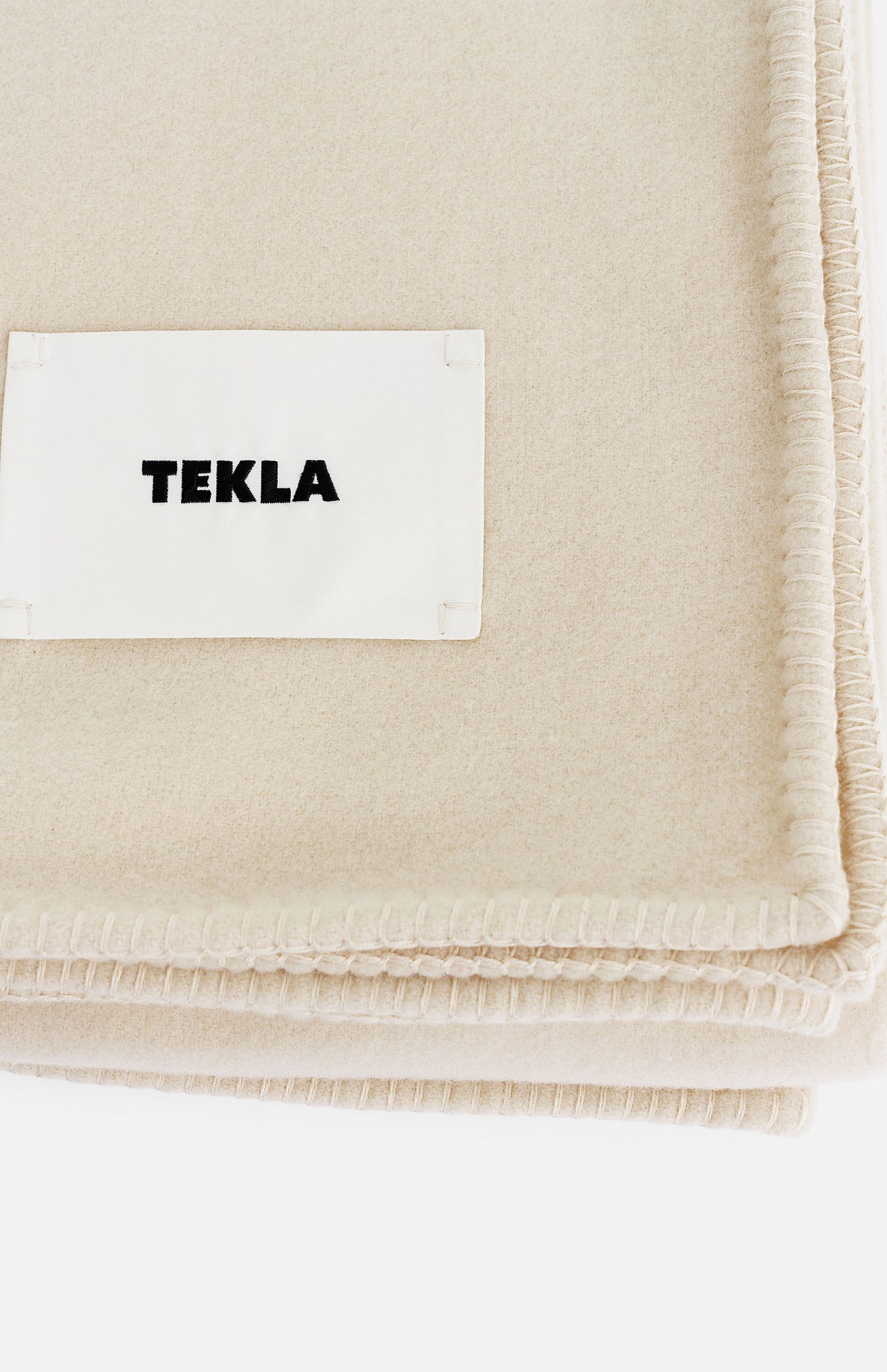 Cream-colored TEKLA Lambswool Blanket with a stitched border and a label.