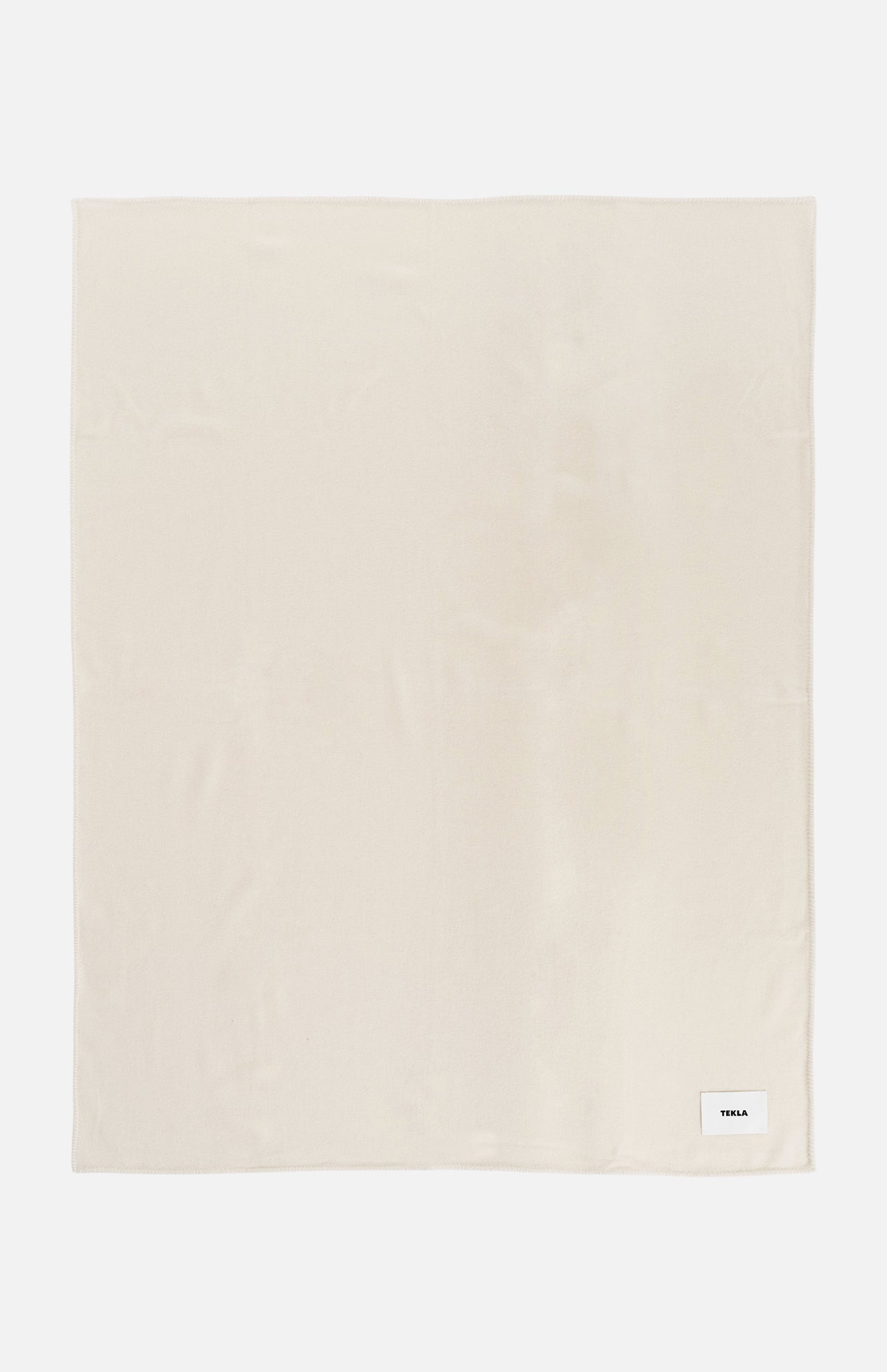Cream-colored rectangular TEKLA Lambswool Blanket with a small woven brand tag in the bottom right corner.
