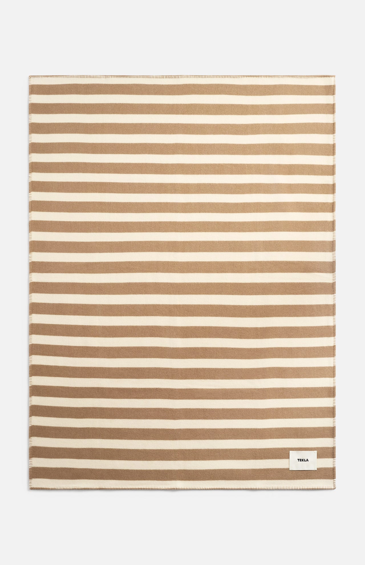 Rectangular TEKLA Striped Lambswool Blanket featuring horizontal tan and cream stripes with a small logo tag in the bottom corner.
