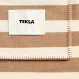 Folded cream and beige striped lambswool blanket by TEKLA with a stitched edge and rectangular fabric tag.