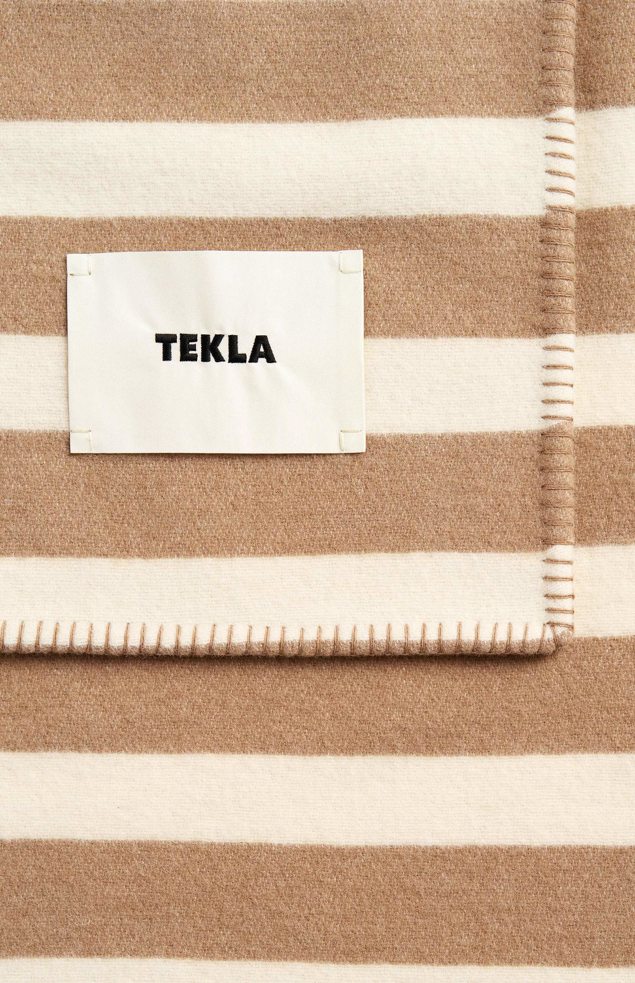 Folded cream and beige striped lambswool blanket by TEKLA with a stitched edge and rectangular fabric tag.