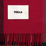 TEKLA Lambswool Fringed Blanket in red, green, and yellow plaid with fringe and a white rectangular brand label stitched in the center.