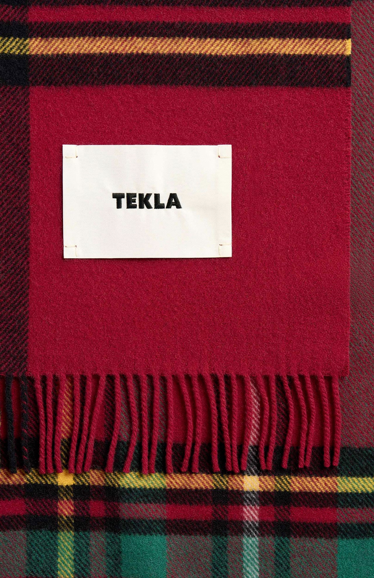 TEKLA Lambswool Fringed Blanket in red, green, and yellow plaid with fringe and a white rectangular brand label stitched in the center.