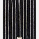 Black rectangular TEKLA Terry Bath Mat made of organic cotton with thin white vertical stripes and a small rectangular brand label at the bottom center.