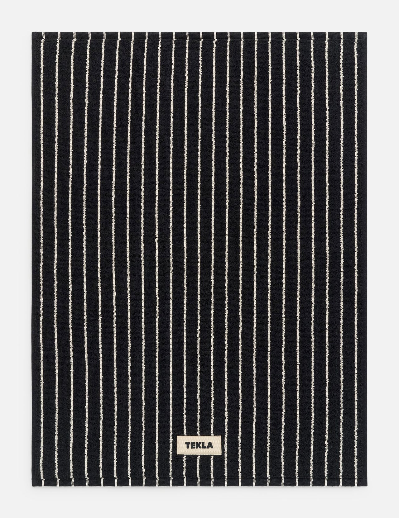 Black rectangular TEKLA Terry Bath Mat made of organic cotton with thin white vertical stripes and a small rectangular brand label at the bottom center.