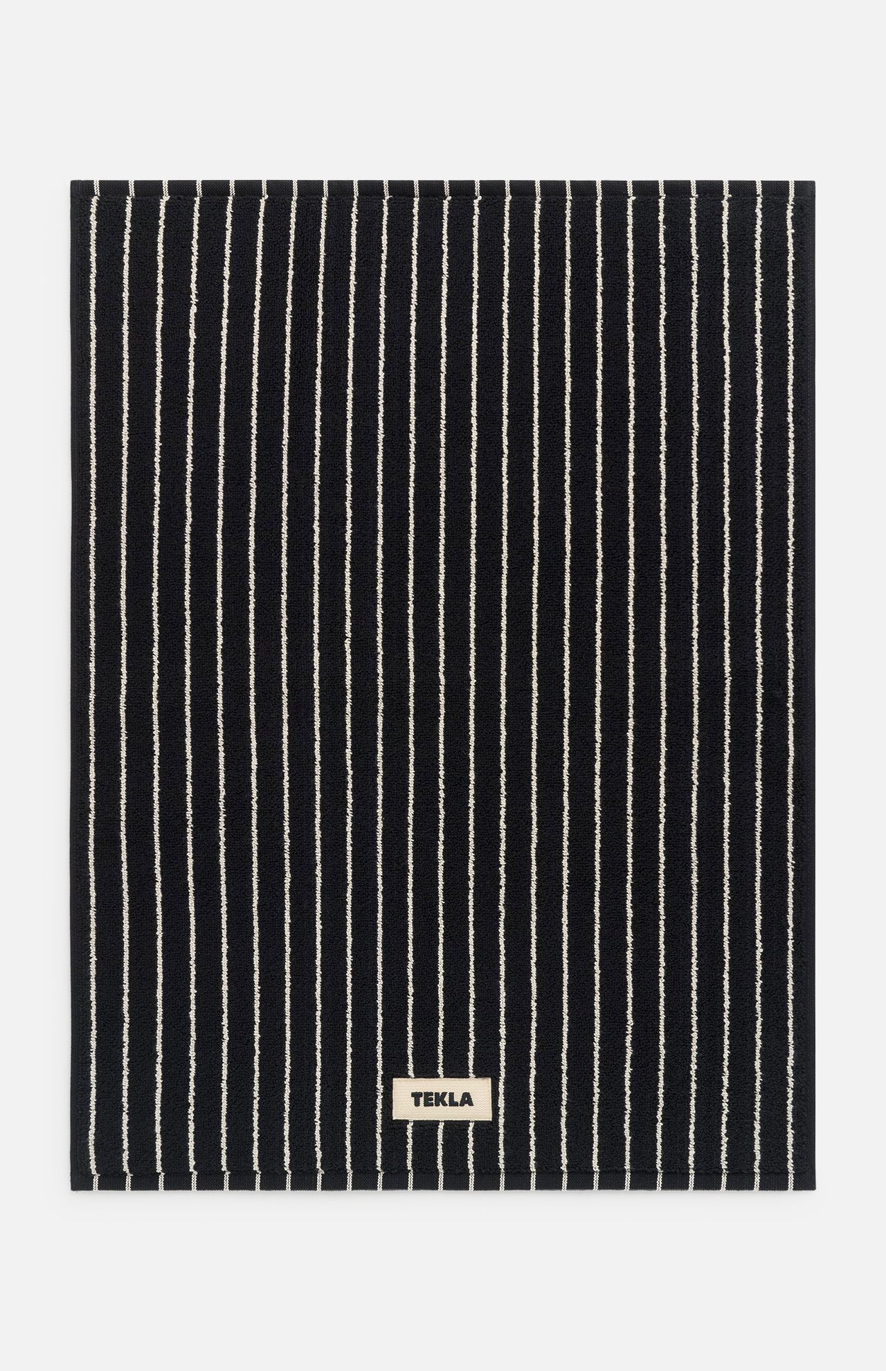 Black rectangular TEKLA Terry Bath Mat made of organic cotton with thin white vertical stripes and a small rectangular brand label at the bottom center.