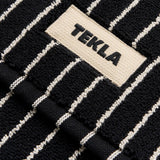 Close-up of a black organic cotton Terry Bath Mat with thin white vertical stripes and a fabric label reading TEKLA.