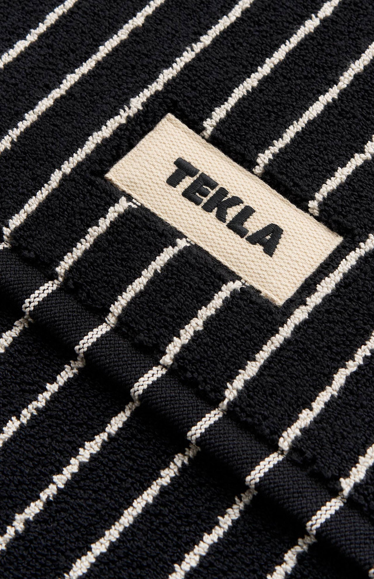 Close-up of a black organic cotton Terry Bath Mat with thin white vertical stripes and a fabric label reading TEKLA.
