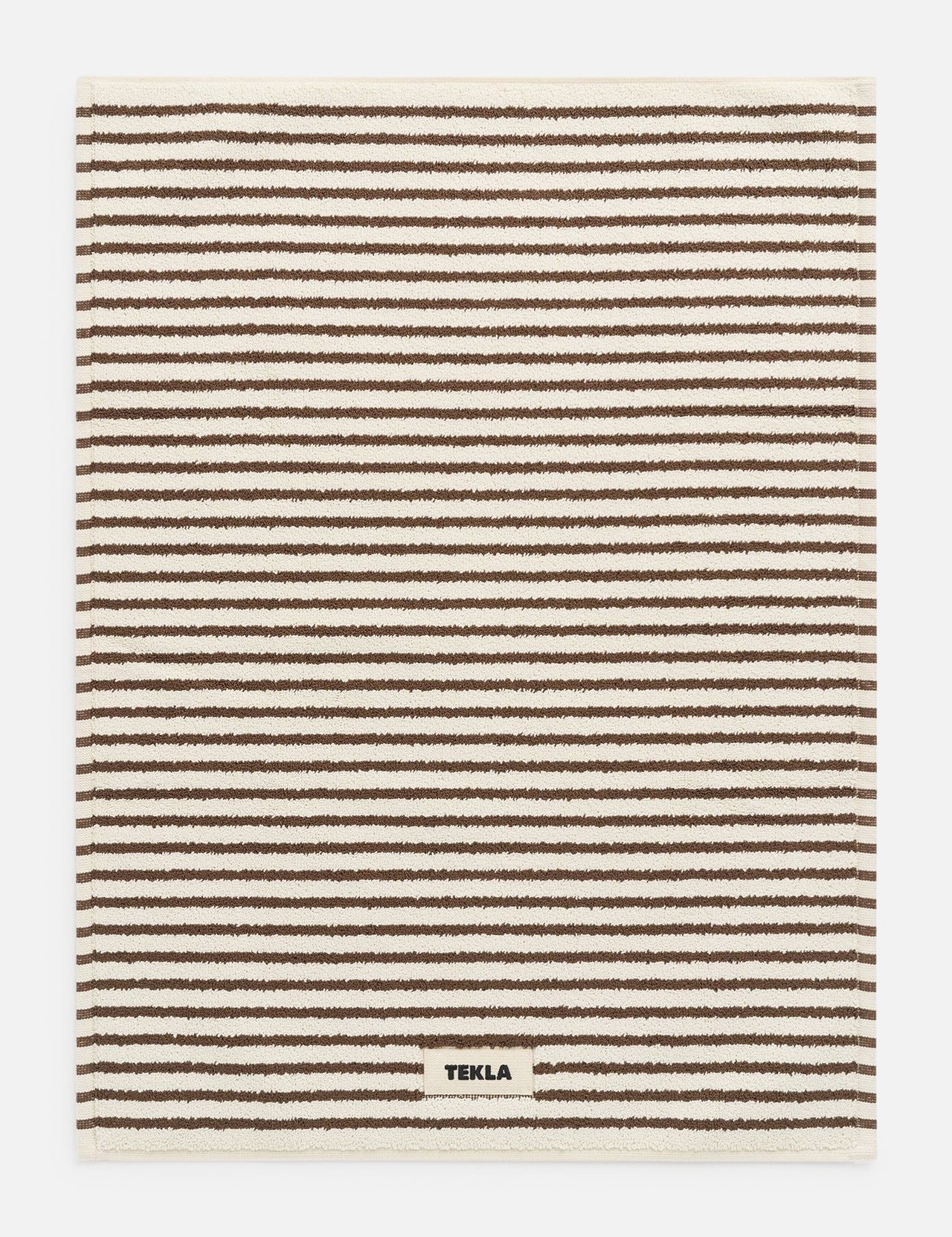Rectangular brown TEKLA Terry Bath Mat with a pattern of thin horizontal brown and cream stripes and a small brand label sewn onto the bottom edge.