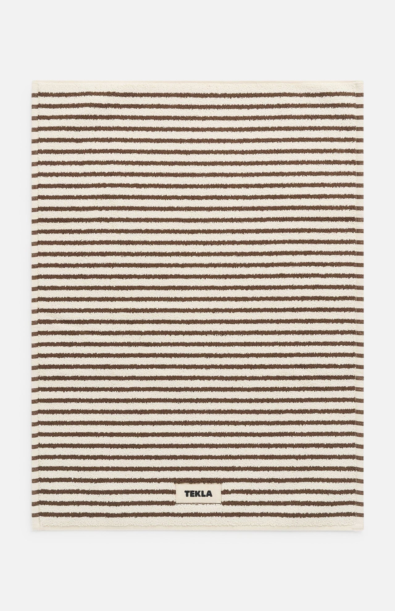 Rectangular brown TEKLA Terry Bath Mat with a pattern of thin horizontal brown and cream stripes and a small brand label sewn onto the bottom edge.