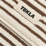 Cream-colored TEKLA Terry Bath Mat in organic cotton with brown stripes and a logo patch.