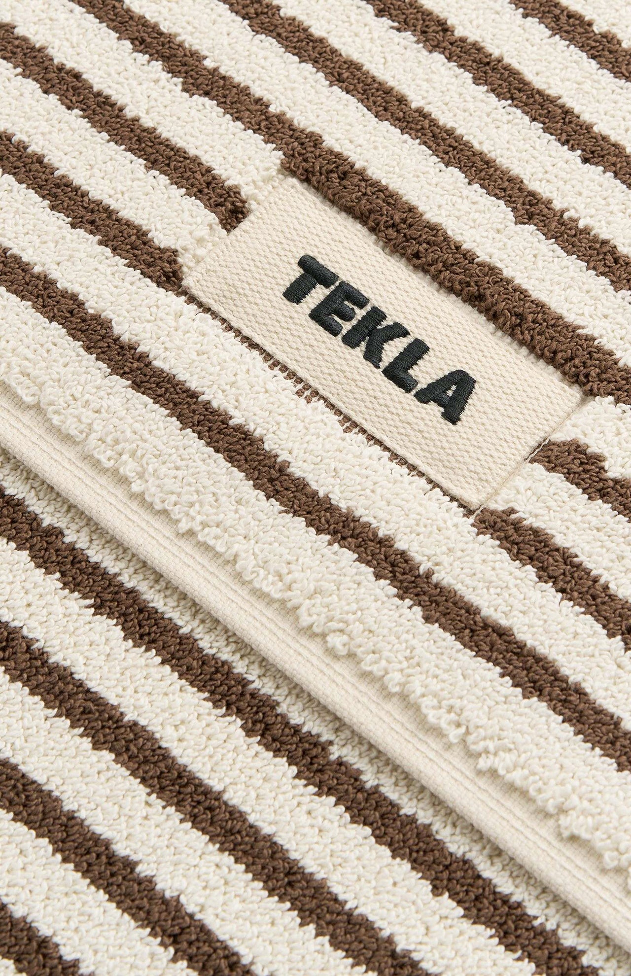 Cream-colored TEKLA Terry Bath Mat in organic cotton with brown stripes and a logo patch.