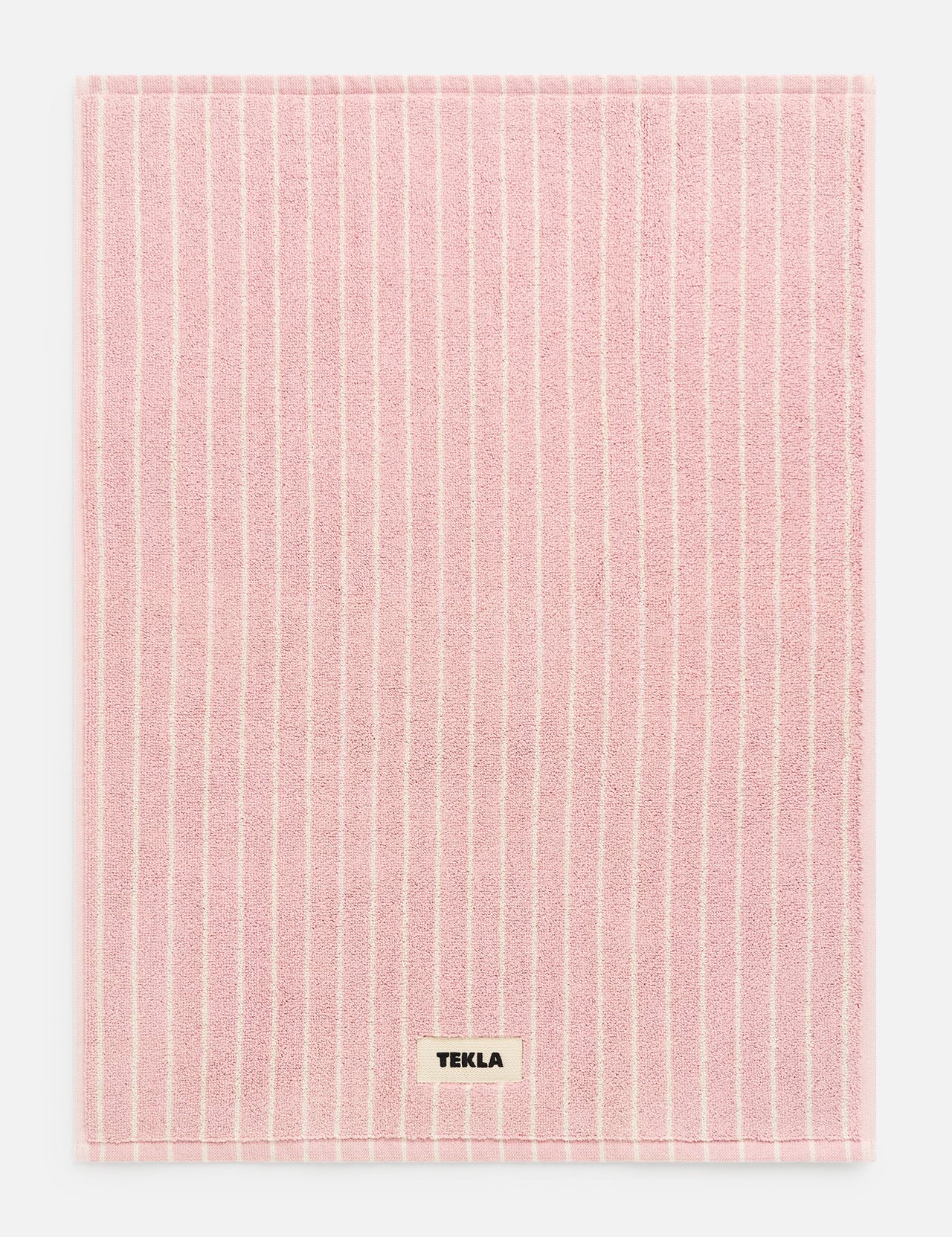 Rectangular pink TEKLA Terry Bath Mat with thin white vertical stripes and a small designer label near the bottom edge.