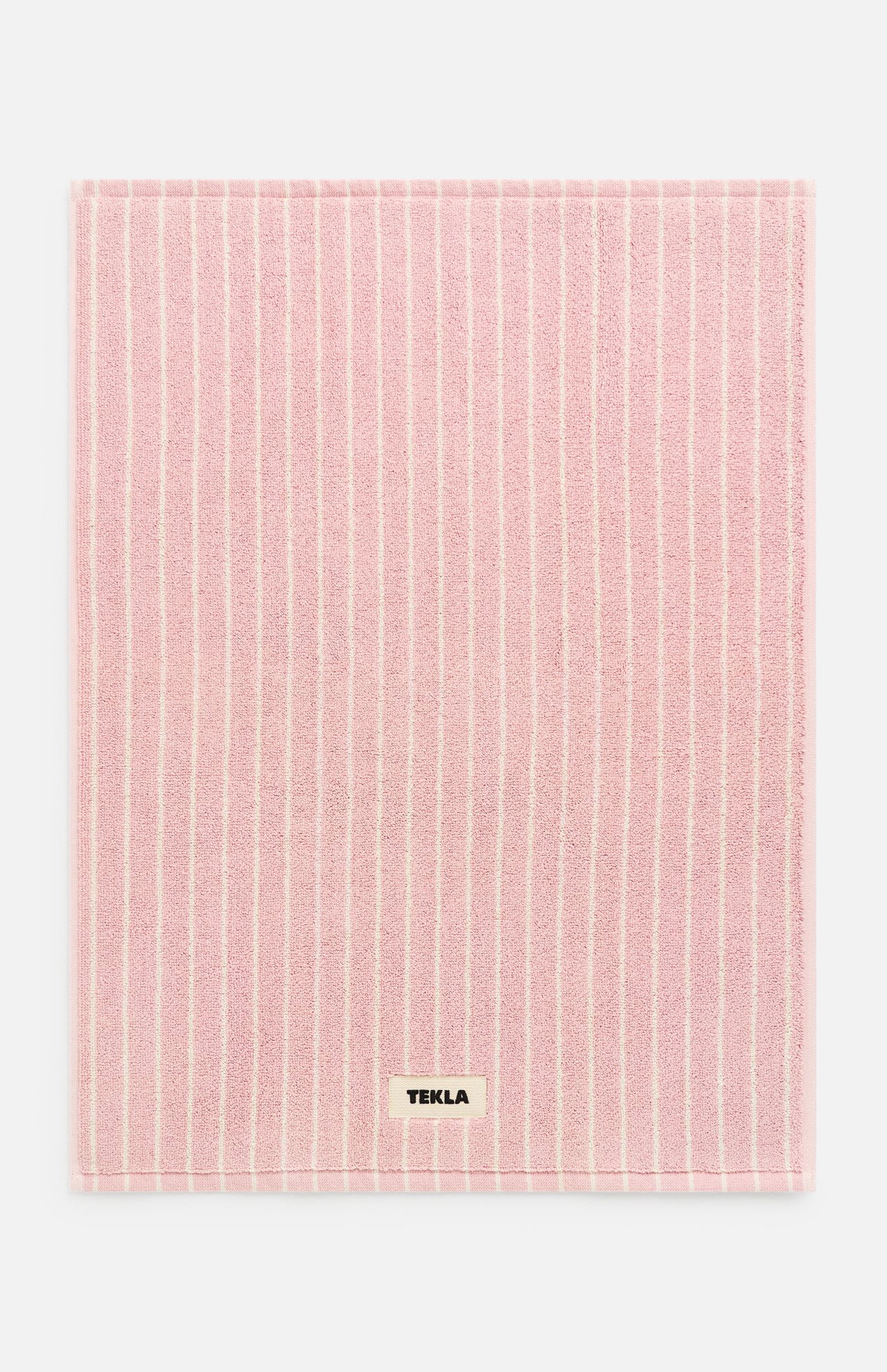 Rectangular pink TEKLA Terry Bath Mat with thin white vertical stripes and a small designer label near the bottom edge.