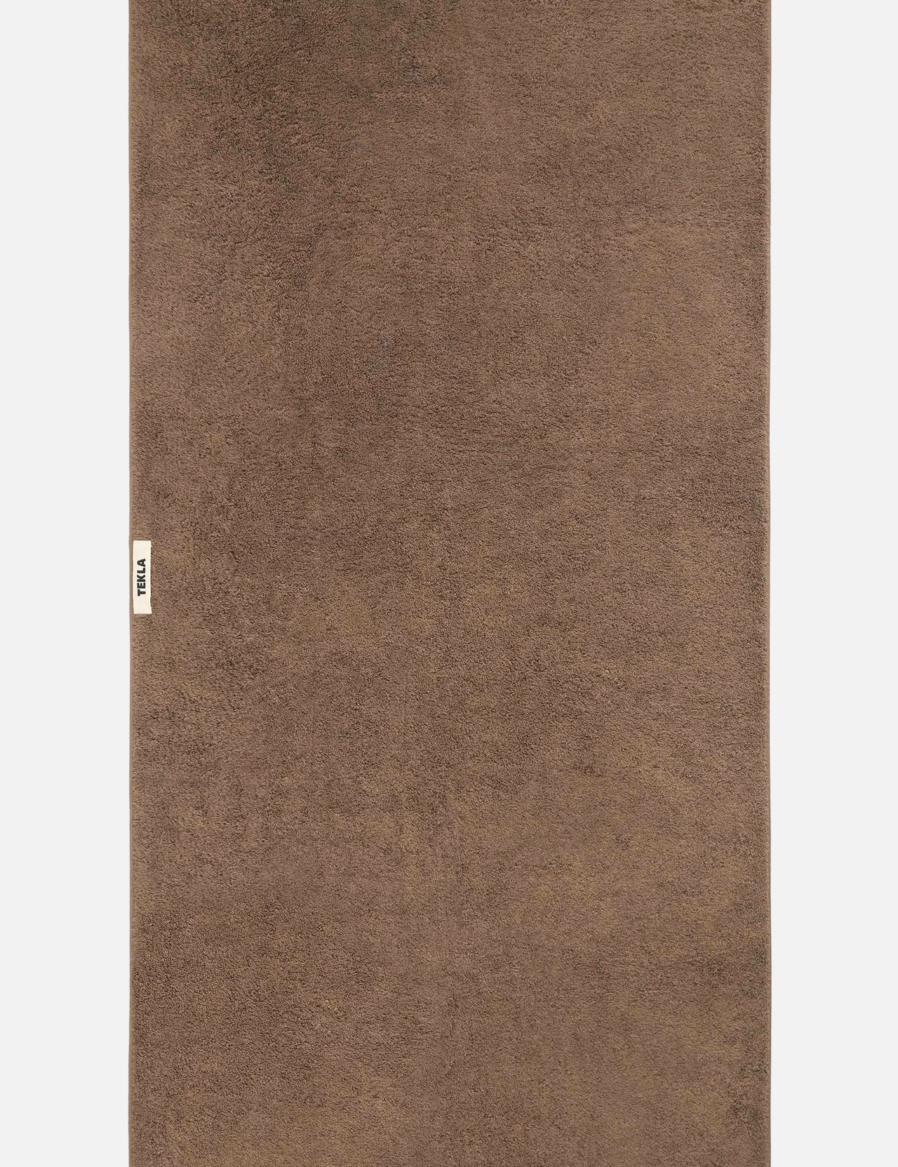 A flat-laid brown organic cotton TEKLA Terry Bath Towel with a small logo tag on the left edge.