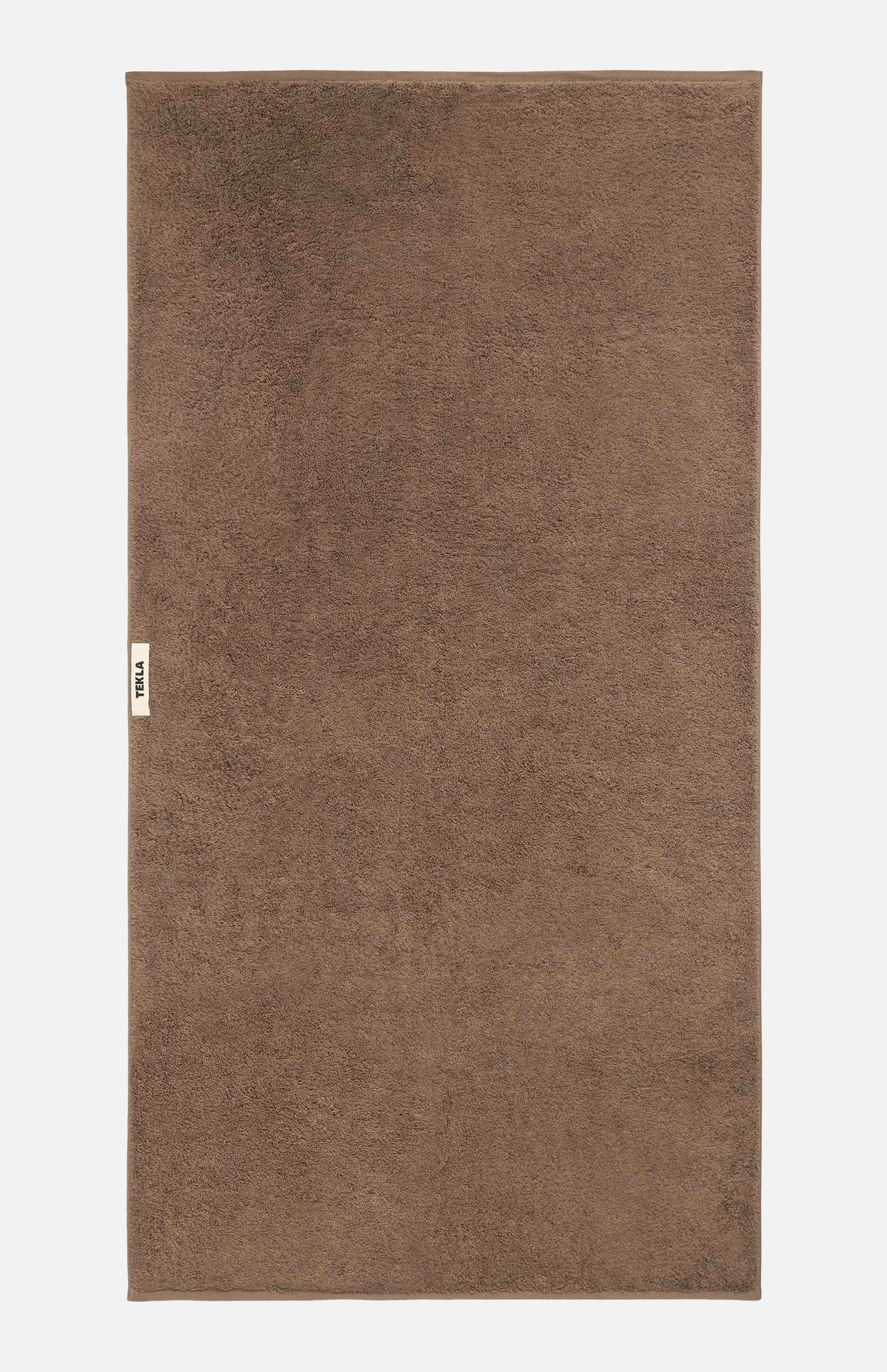 A flat-laid brown organic cotton TEKLA Terry Bath Towel with a small logo tag on the left edge.