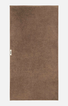 A flat-laid brown organic cotton TEKLA Terry Bath Towel with a small logo tag on the left edge.