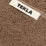 Close-up of a brown organic cotton Terry Bath Towel by TEKLA, featuring a white label with the brand name in black.