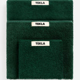 Three folded forest green TEKLA Terry Bath towels stacked, each with a cream-colored brand tag centered at the top edge.