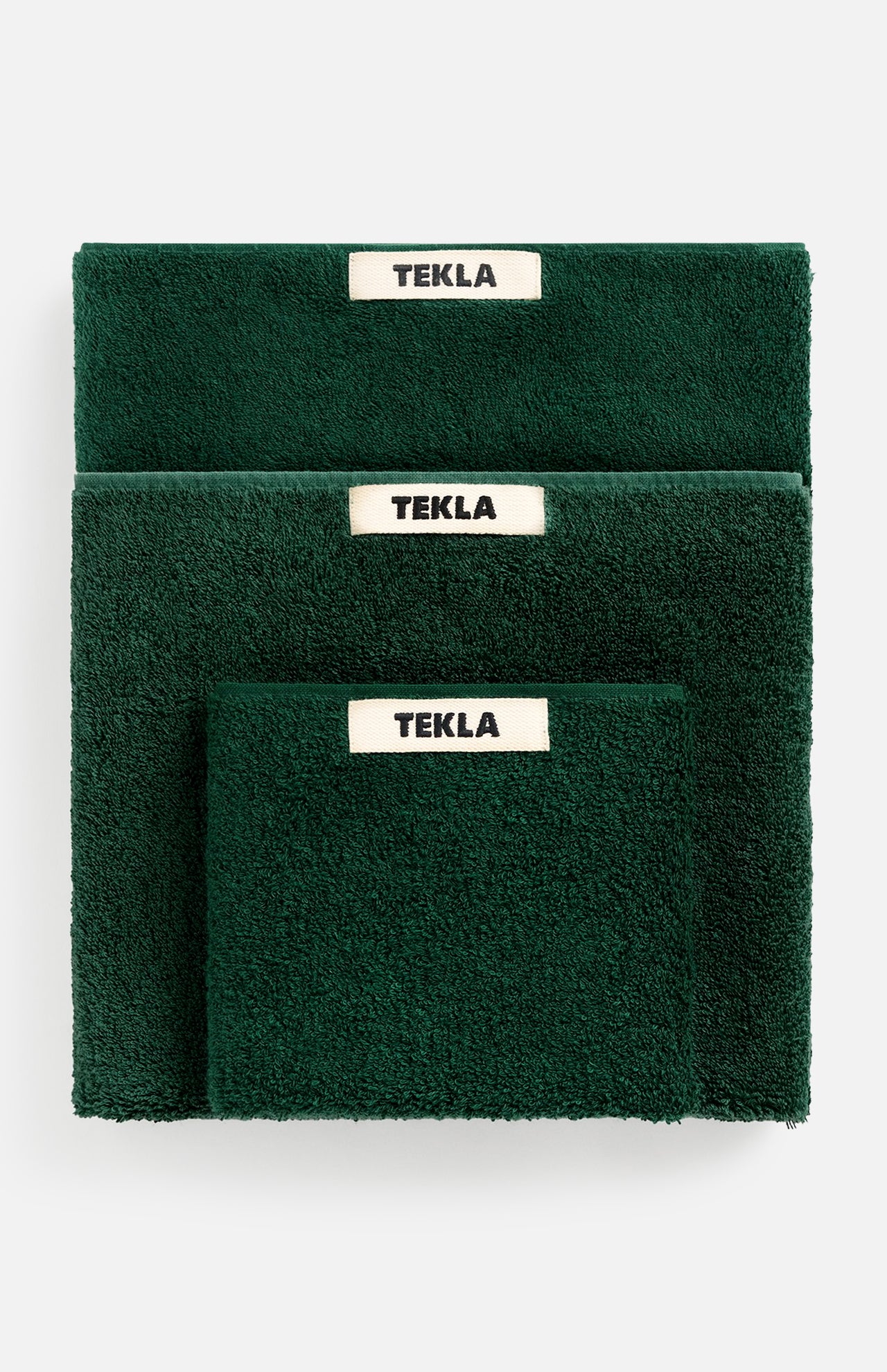 Three folded forest green TEKLA Terry Bath towels stacked, each with a cream-colored brand tag centered at the top edge.