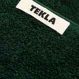Close-up of a forest green Terry Bath Towel with a white rectangular fabric label that reads TEKLA in black text.