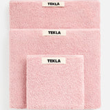 Three stacked pink TEKLA Terry Bath Towels of varying sizes, each with a central fabric tag.