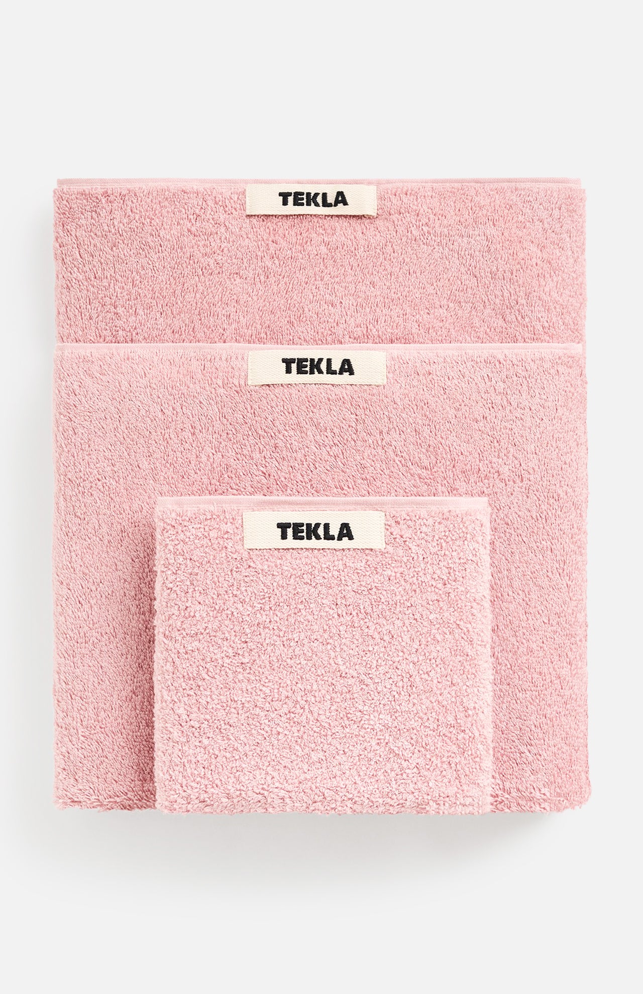 Three stacked pink TEKLA Terry Bath Towels of varying sizes, each with a central fabric tag.