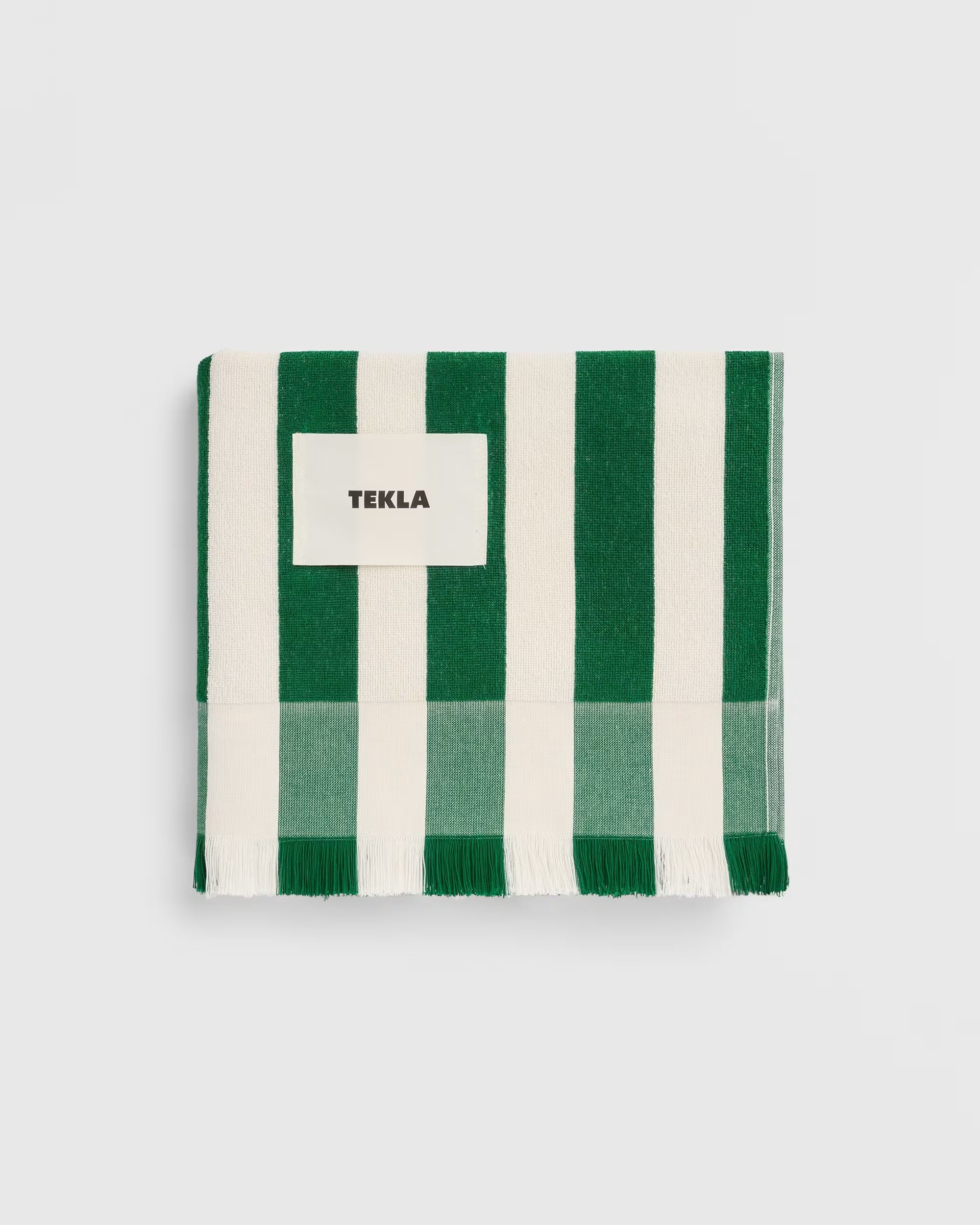 Folded dark green and cream striped TEKLA Terry Beach Towel with fringe and a central logo patch.