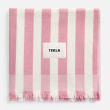 A pink and white striped TEKLA Terry Beach Towel made of organic cotton with a fringed edge and a central label.