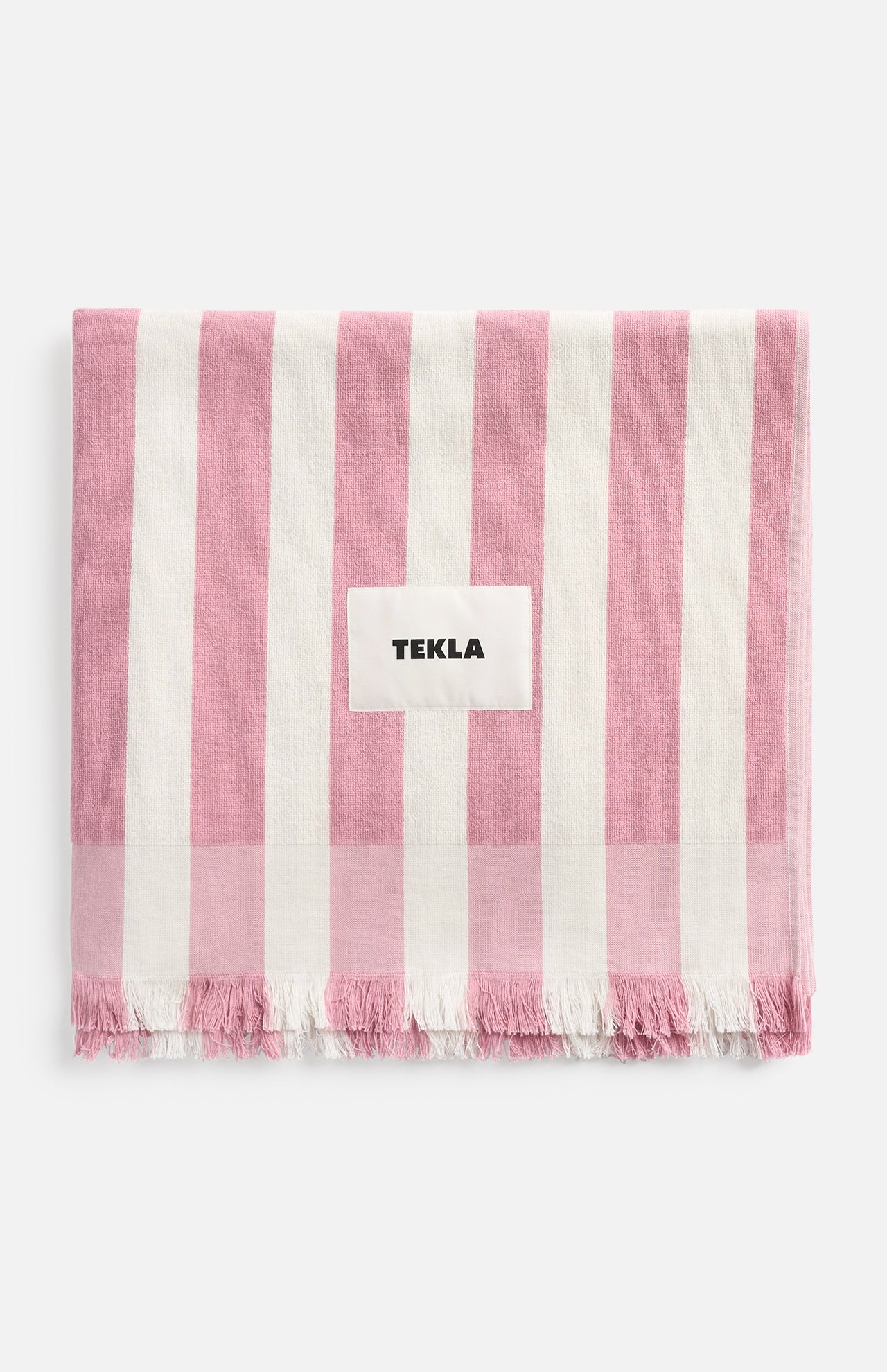 A pink and white striped TEKLA Terry Beach Towel made of organic cotton with a fringed edge and a central label.