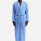 A person wearing a TEKLA Terry Classic Unisex Bathrobe in blue organic cotton with white vertical pinstripes, tied at the waist with a matching belt.