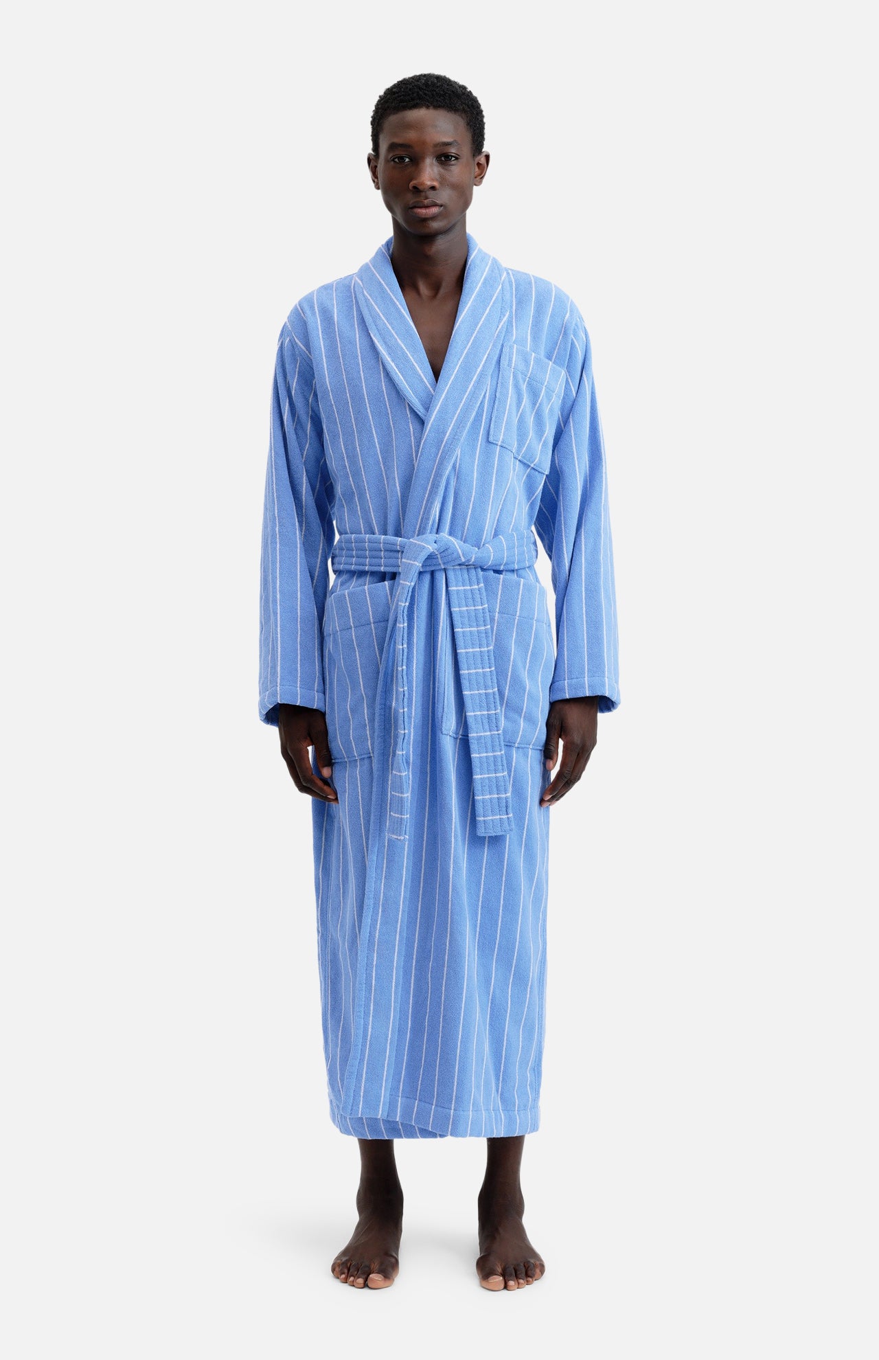 A person wearing a TEKLA Terry Classic Unisex Bathrobe in blue organic cotton with white vertical pinstripes, tied at the waist with a matching belt.