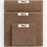 Three brown organic cotton TEKLA Terry Hand Towels of varying sizes stacked on top of one another, each featuring a white rectangular brand label.