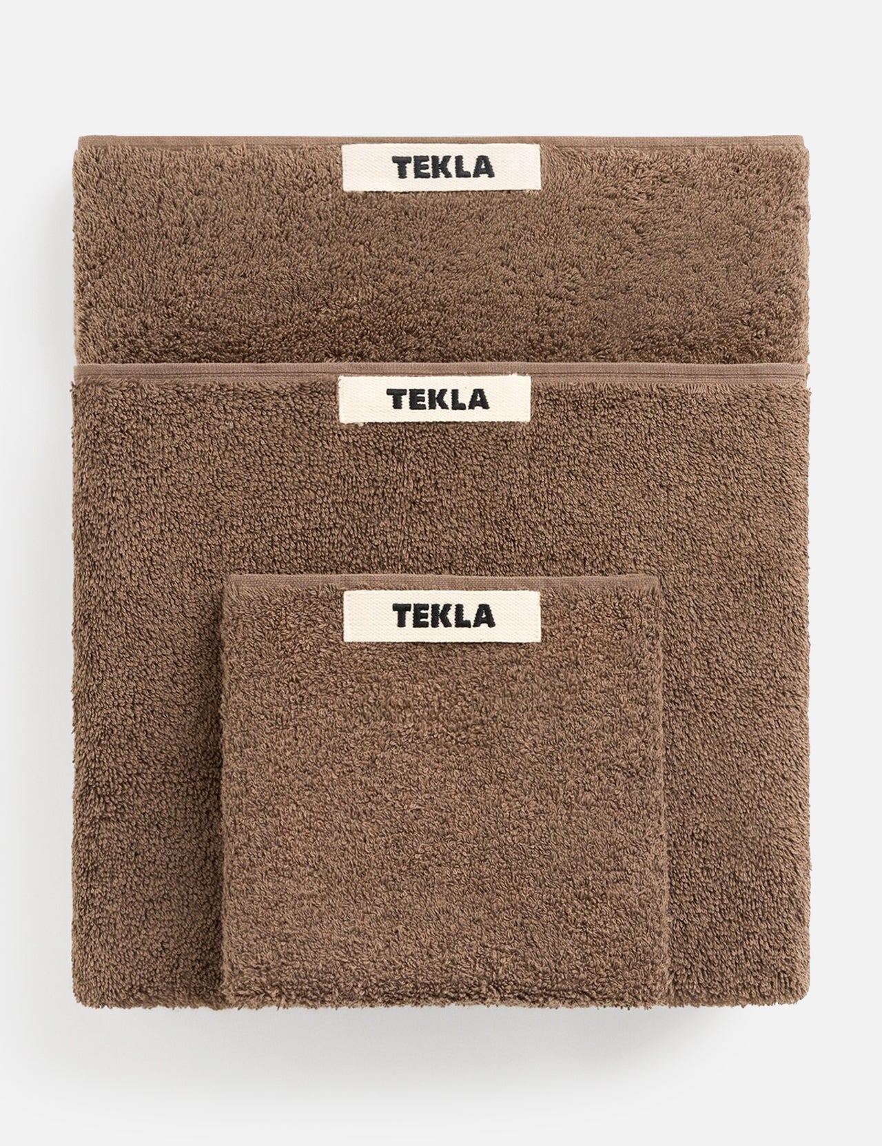 Three brown organic cotton TEKLA Terry Hand Towels of varying sizes stacked on top of one another, each featuring a white rectangular brand label.