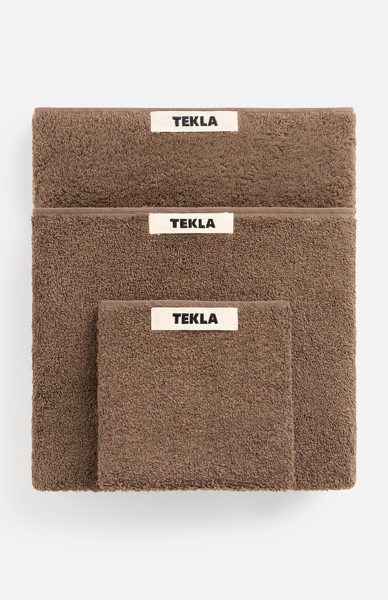 Three brown organic cotton TEKLA Terry Hand Towels of varying sizes stacked on top of one another, each featuring a white rectangular brand label.