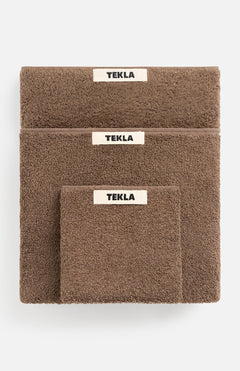 Three brown organic cotton TEKLA Terry Hand Towels of varying sizes stacked on top of one another, each featuring a white rectangular brand label.
