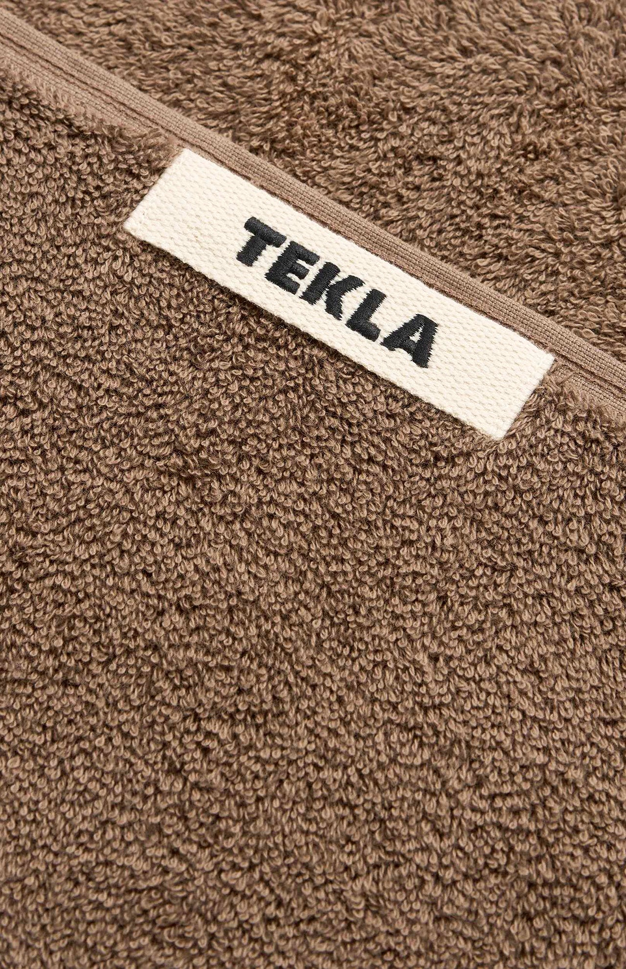 Close-up of a brown organic cotton Terry Hand Towel with a white rectangular label featuring the text TEKLA in black.