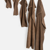 Five brown organic cotton TEKLA Terry Hand Towels of varying sizes hanging in a row against a white wall.