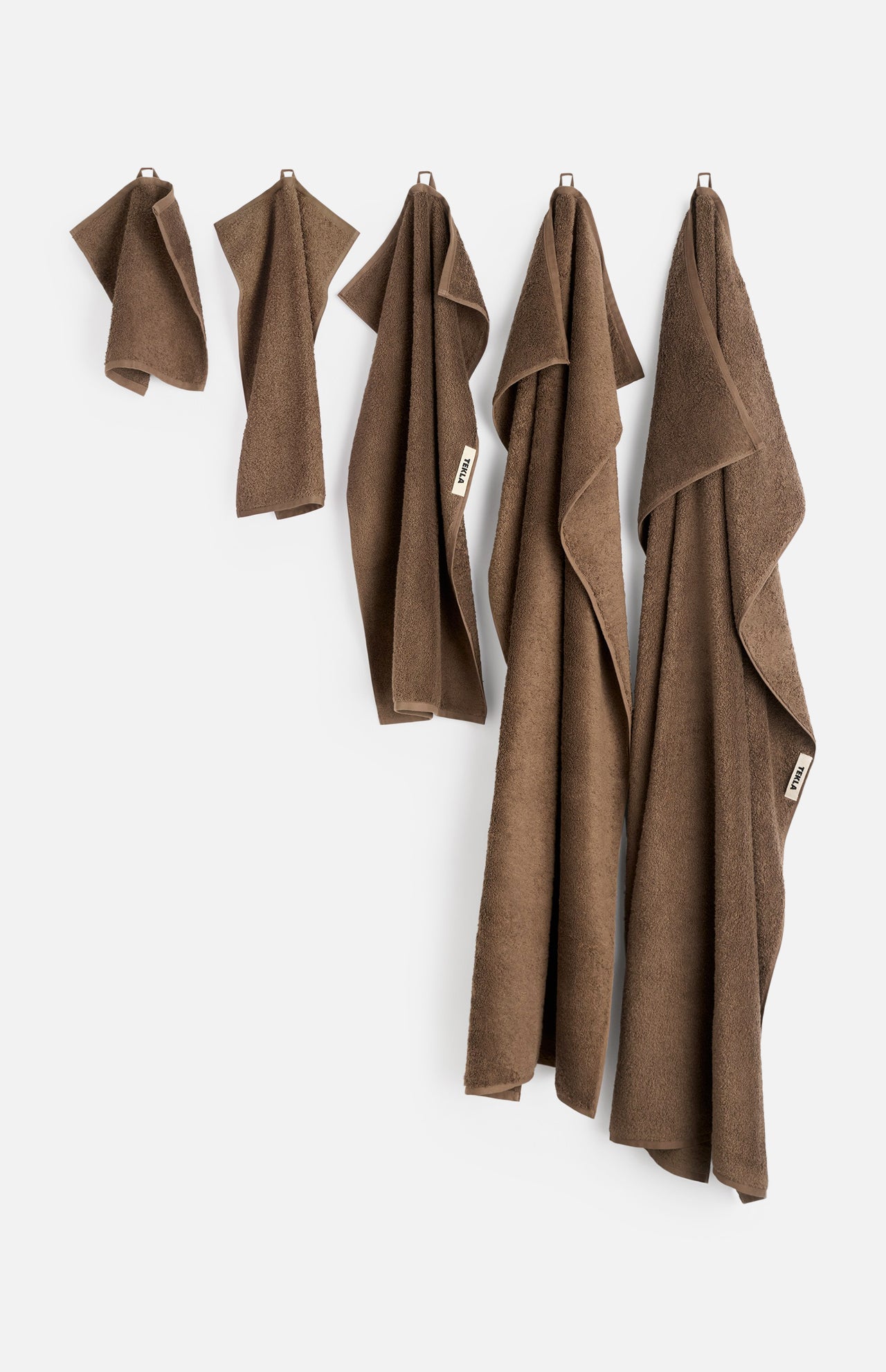 Five brown organic cotton TEKLA Terry Hand Towels of varying sizes hanging in a row against a white wall.