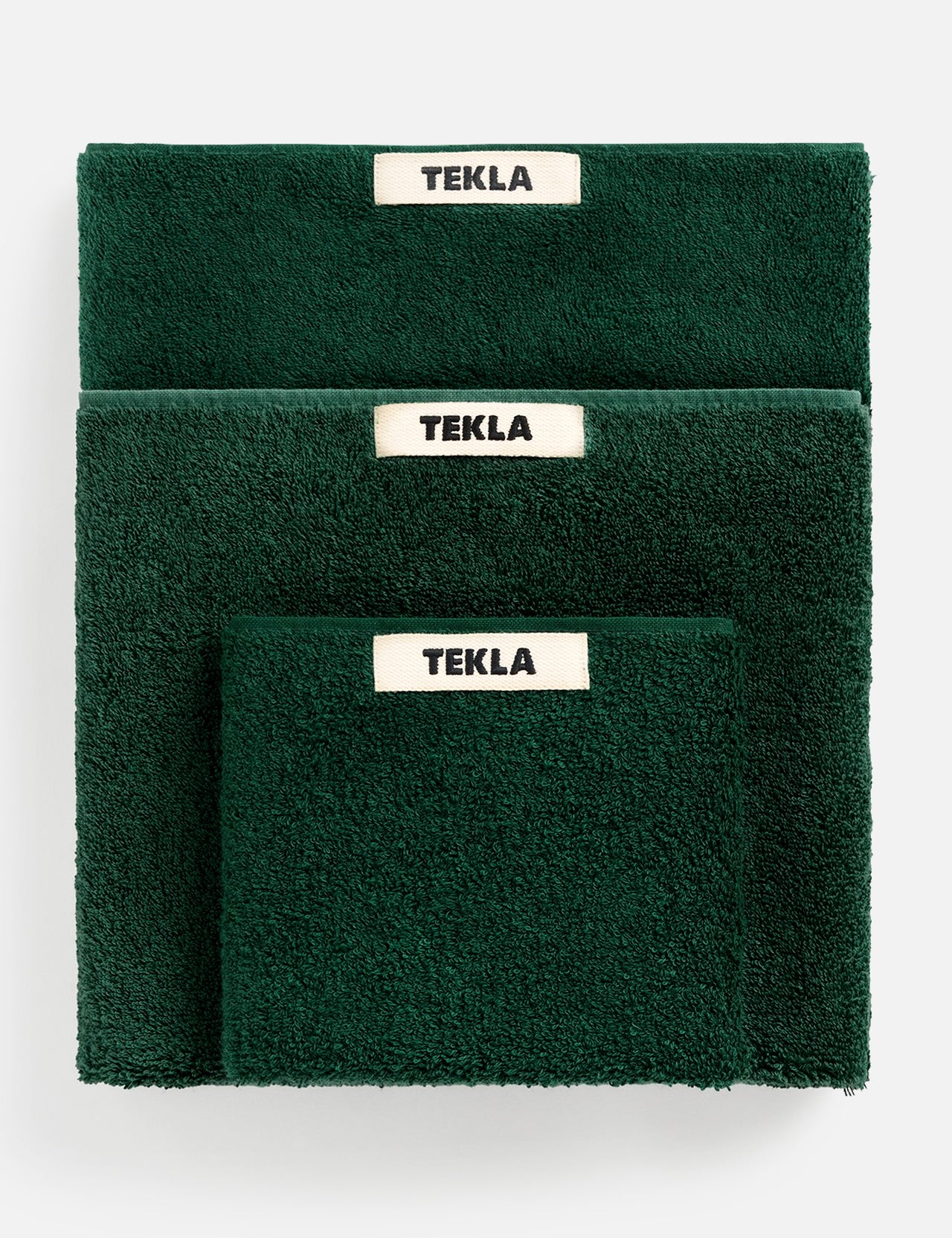 Three forest green TEKLA Terry Hand Towels of varying sizes stacked together, each featuring a white label with black text.