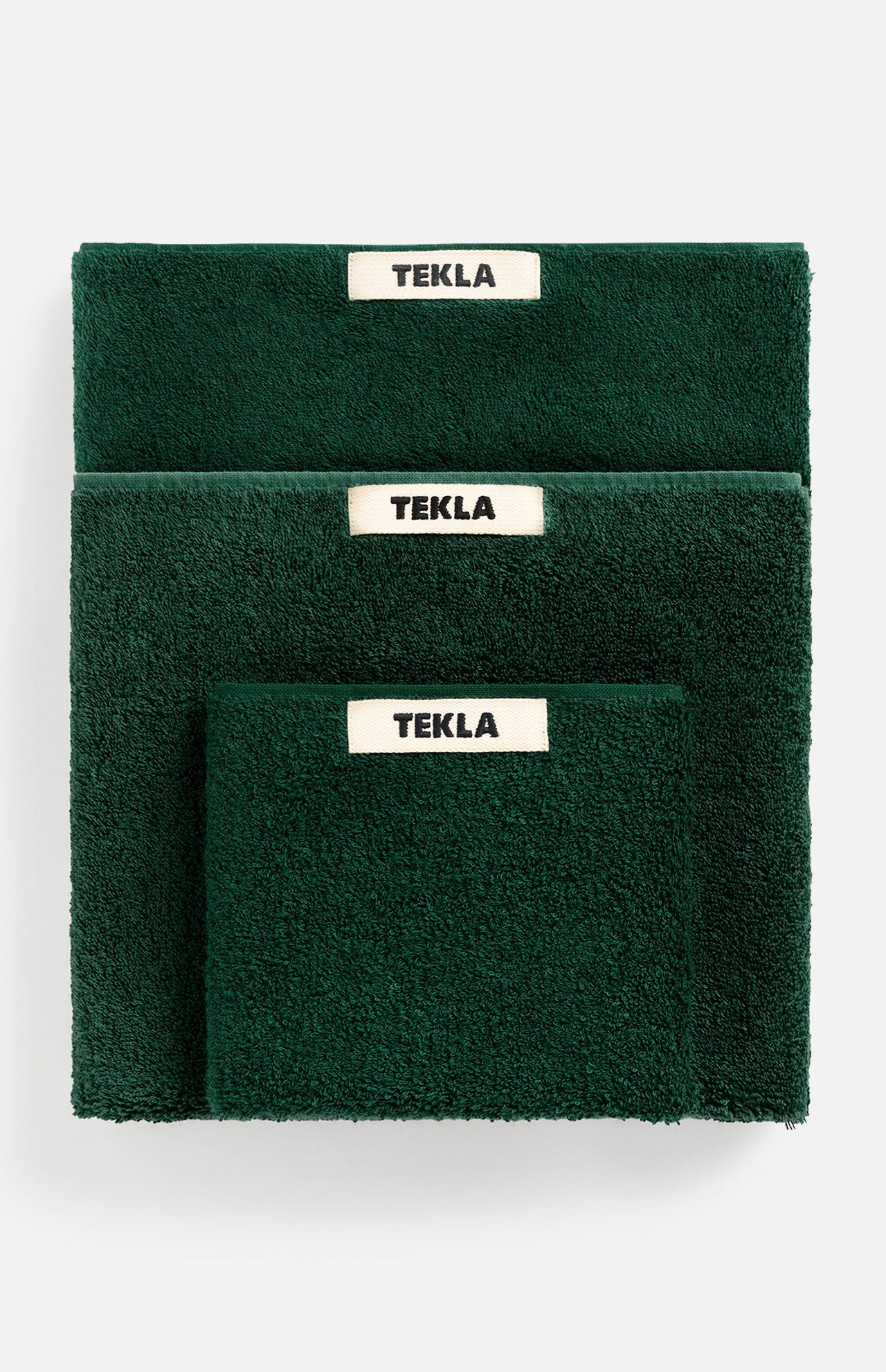 Three forest green TEKLA Terry Hand Towels of varying sizes stacked together, each featuring a white label with black text.
