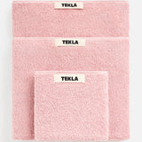 Three folded pink TEKLA Terry Hand Towels of varying sizes stacked on top of each other, each featuring a centered brand label.