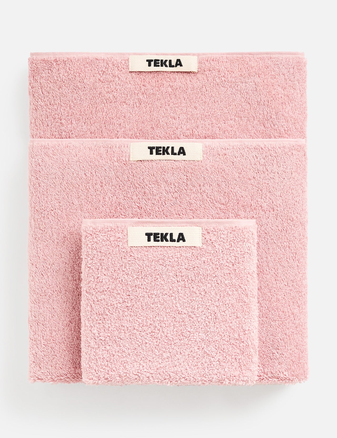 Three folded pink TEKLA Terry Hand Towels of varying sizes stacked on top of each other, each featuring a centered brand label.